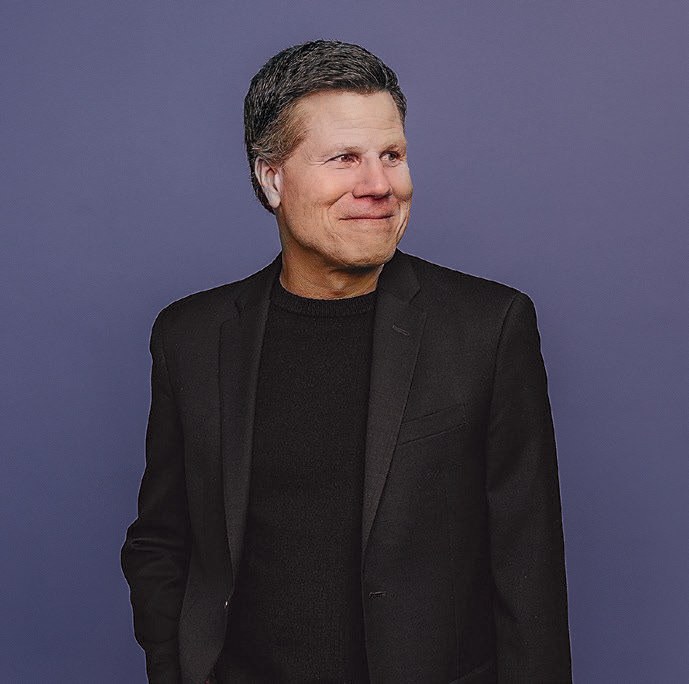 A Man in a black shirt and jacket with gray hair looks off to the right and smiles