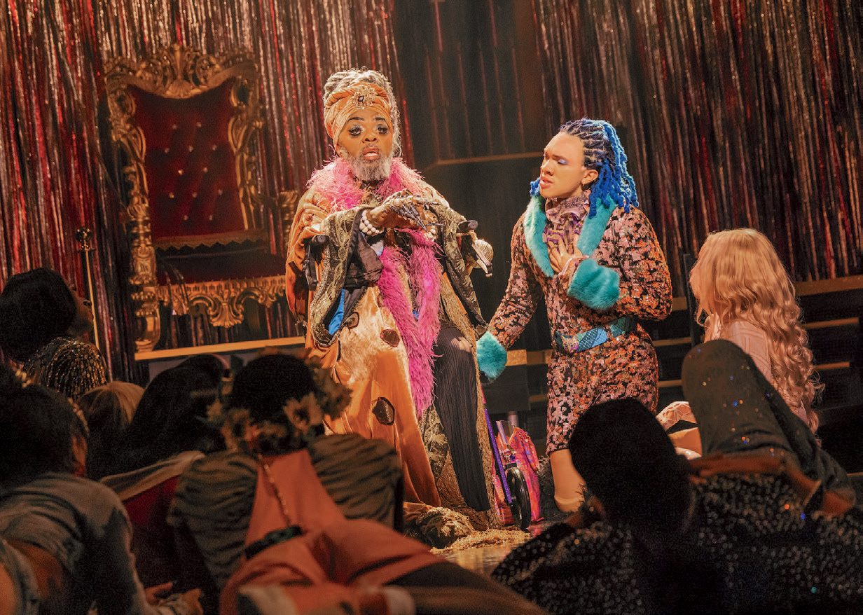 Performers wearing colorful costumes stand in front of a red curtain and throne and speak to the audience