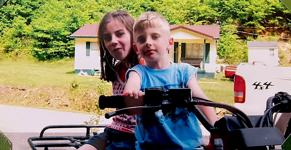 A young girl and bow sitting on a four-wheeler in front of a house and trees