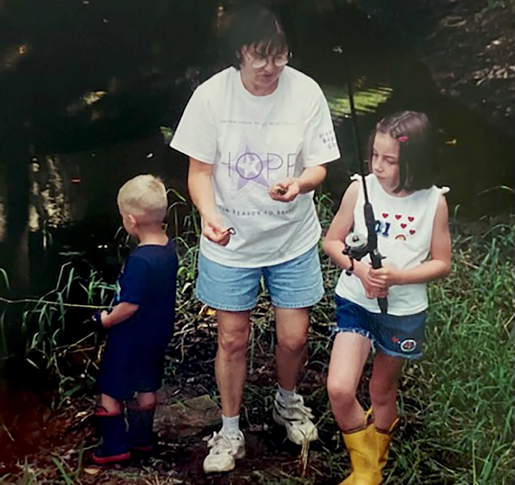 A woman with glasses and a white t shirt talking to a young girl holding a fishing rod, a young boy in a navy t shirt turns away from the camera and stands next to them