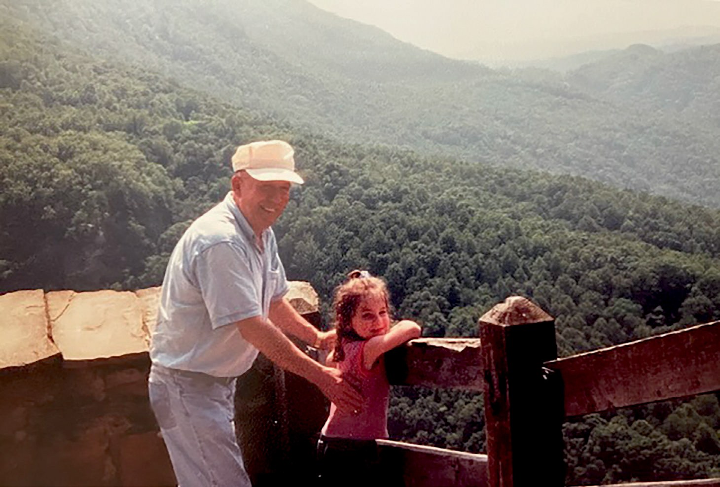 an older man wearing a white cap holds a young girl while she leans over a wooden fence and smiles, in front of a landscape of green mountains