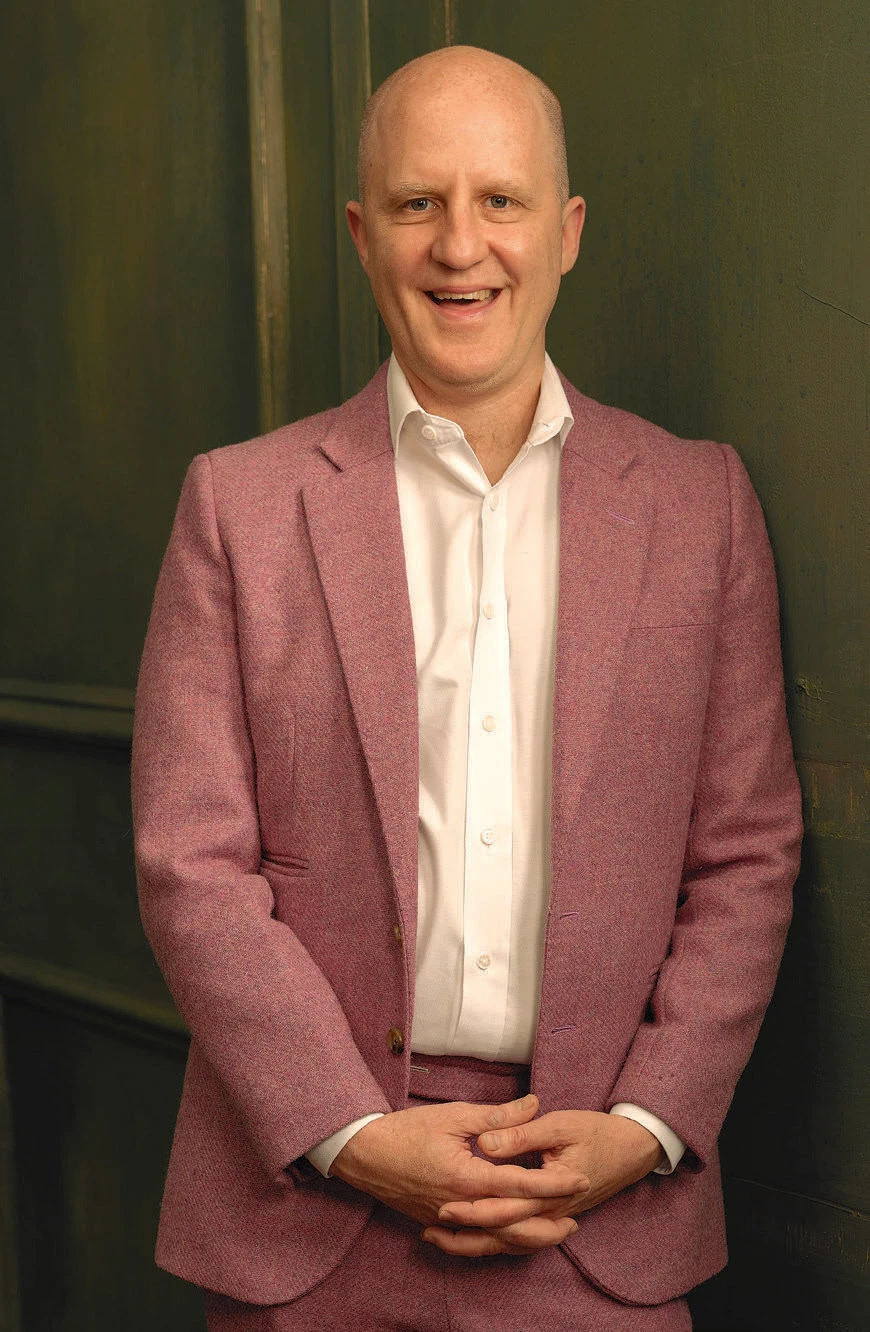 Director Sandi DuBowski smiling in pink suit against dark green wall.