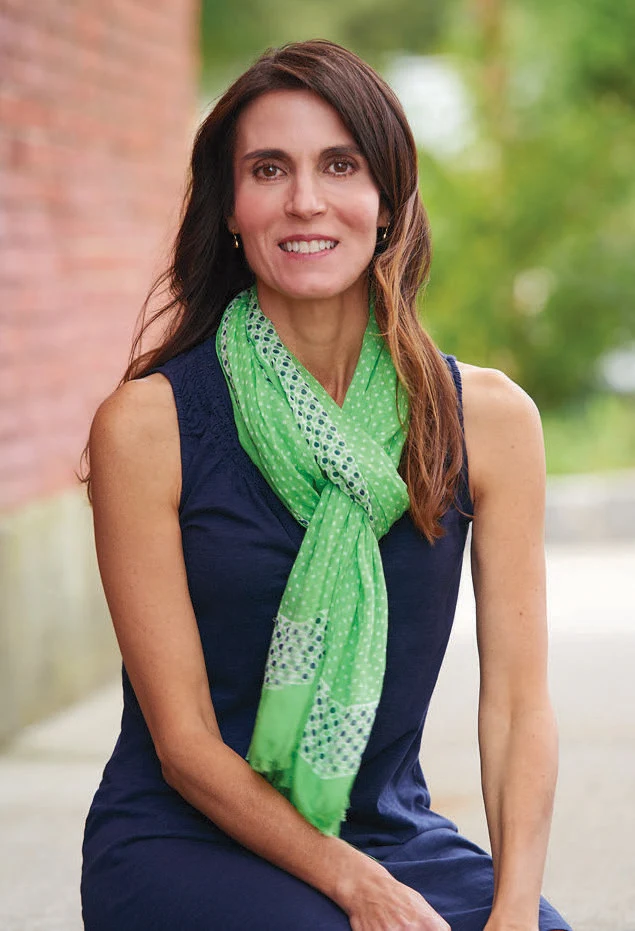 A woman miling wearing a green scarf in front of a blurred background with a brick wall and trees