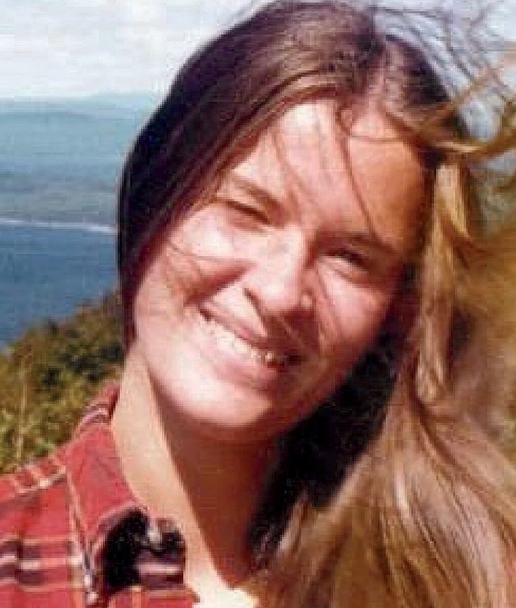A woman smiling wearing a red plaid shirt, in front of a body of water
