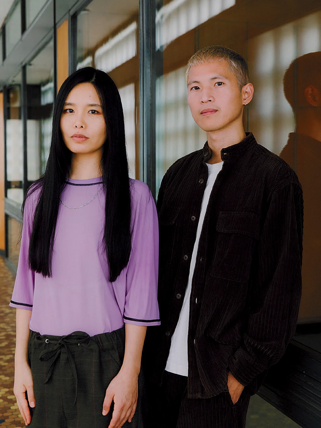 A woman with long black hair and a purple shirt standing to the left of a man with short blond hair and a black jacket, both with neutral expressions