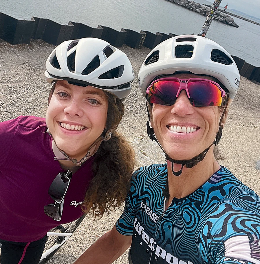 Two women smiling wearing bike helmets in front of a body of water