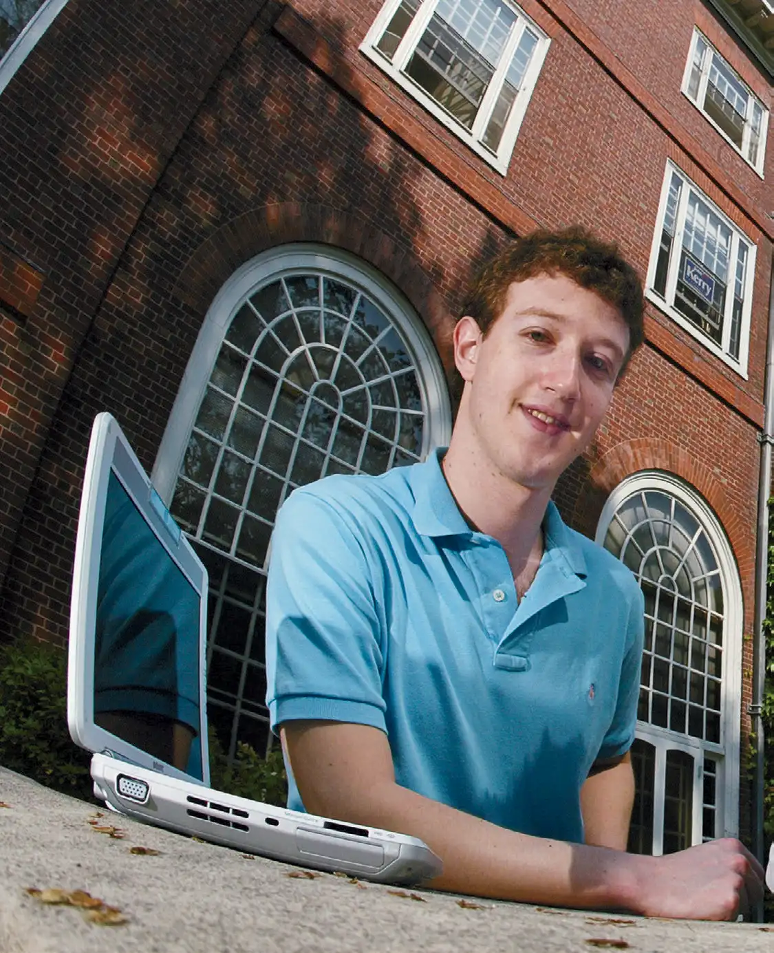 A man in a blue polo sitting behind an open retro silver laptop in front of a brick building