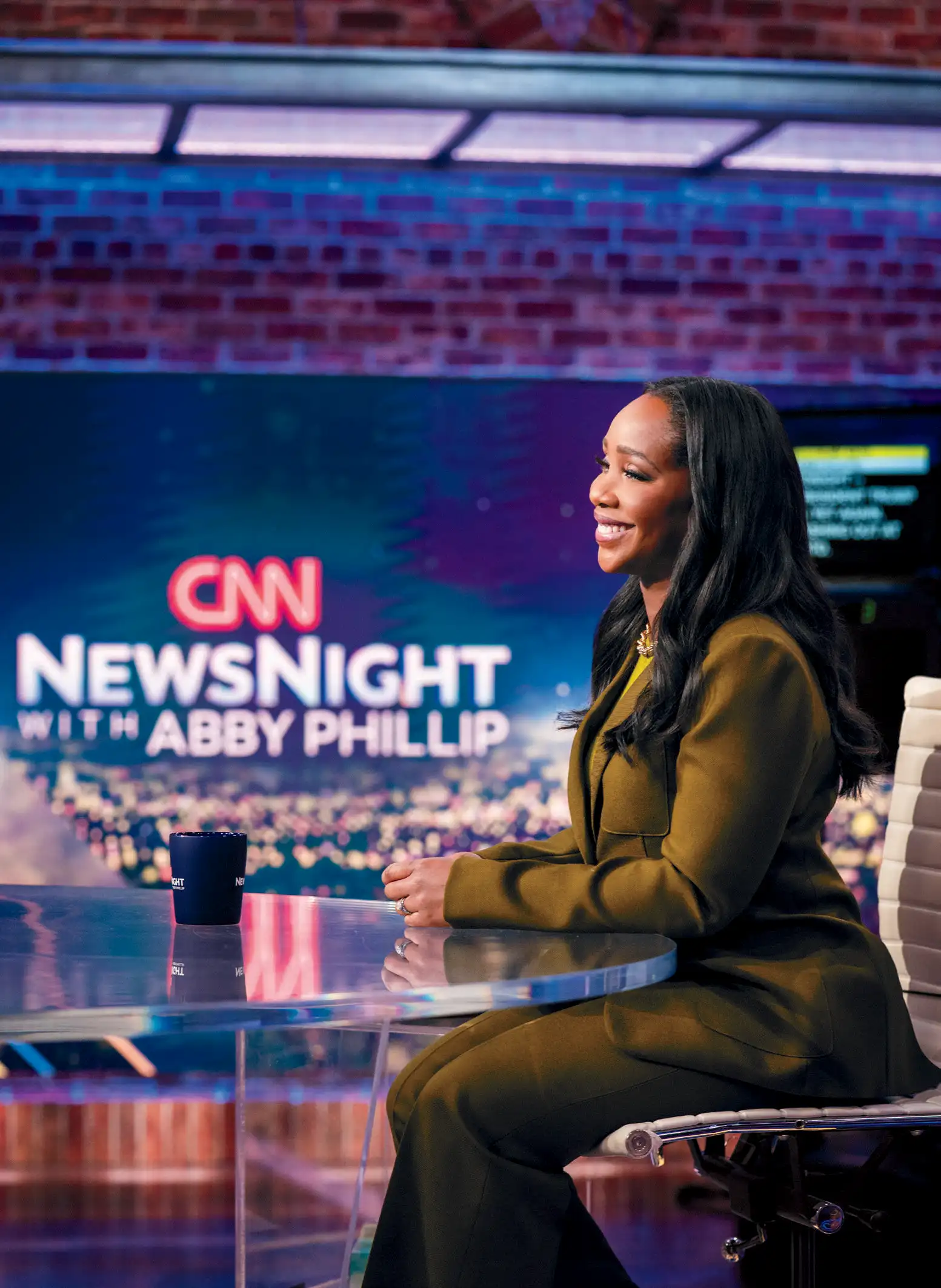 A woman in a blazer sits at a table smiling in front of a backdrop that says "CNN NewsNight with Abby Phillip"