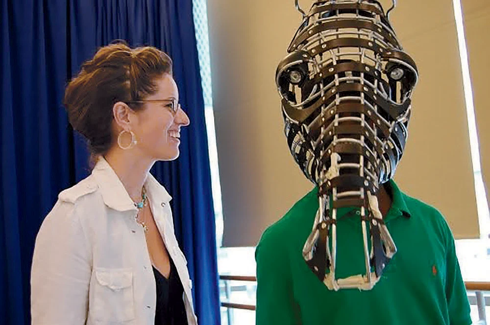 A woman in a white jacket smiles at a man wearing a wooden mask of a horse