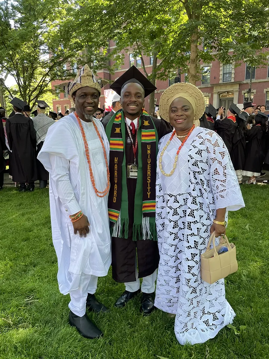 Three people smiling: In the center, a man wearing black mortarboard cap, black graduation gown, with black, green, yellow, and red stole that reads "Harvard University." To his left, a man in whitee gown and yellow hat and orange beaded necklace. To his right, a woman wearing a round yellow hat, white gown, and orange and yellow beaded necklace and holding a tan purse