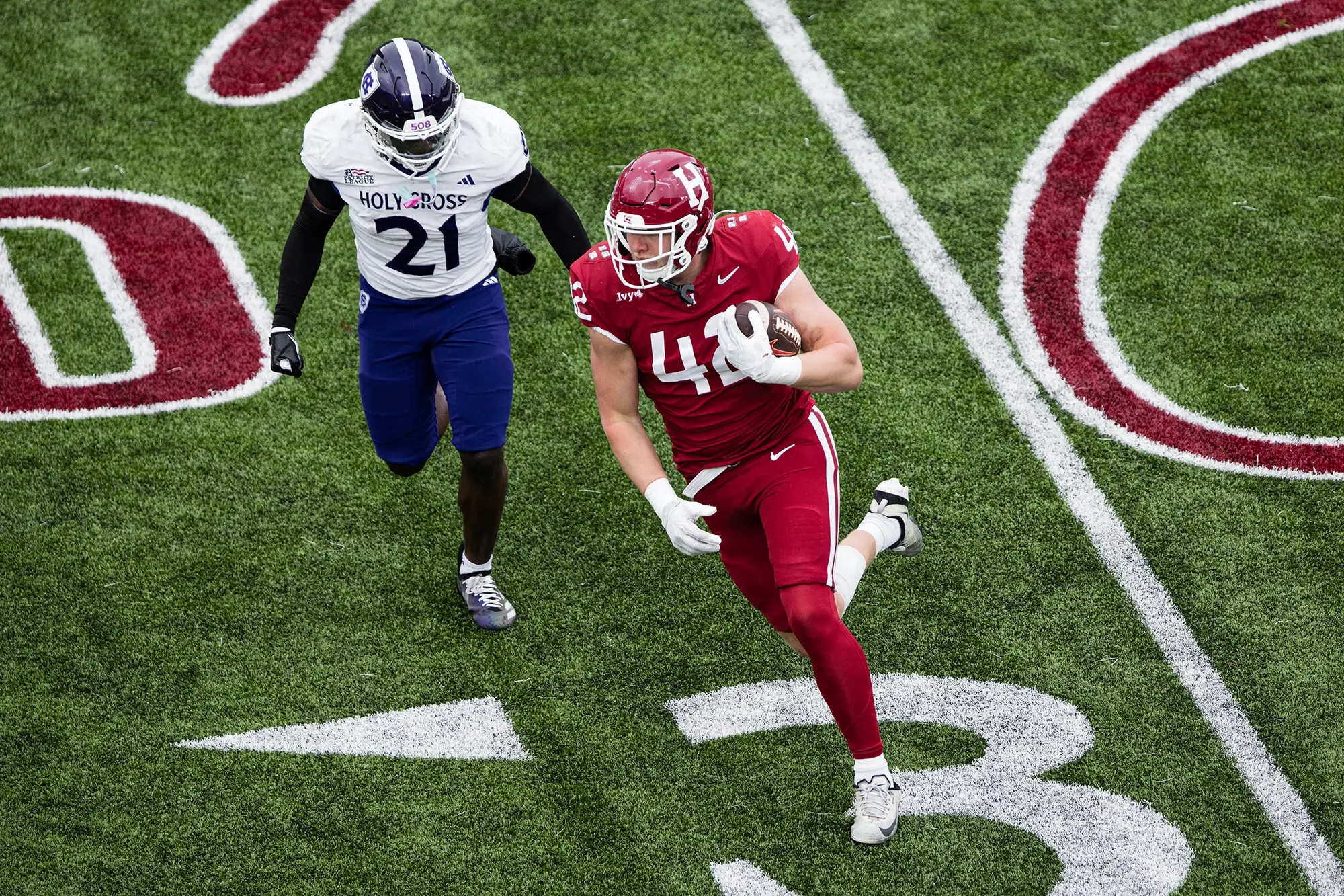 Harvard player #42, wearing a red uniform, runs with the ball in hand as Holy Cross player #21, wearing white and blue uniform, tries to intercept