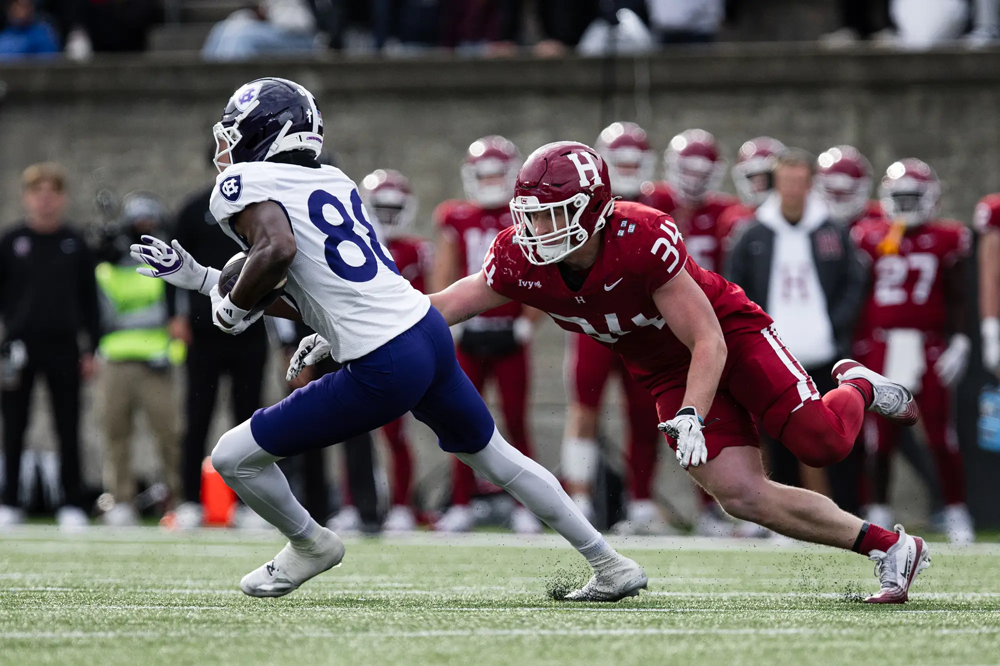 Harvard player #34, wearing a red uniform, dives at Holy Cross player, wearing white and blue, who is holding the ball