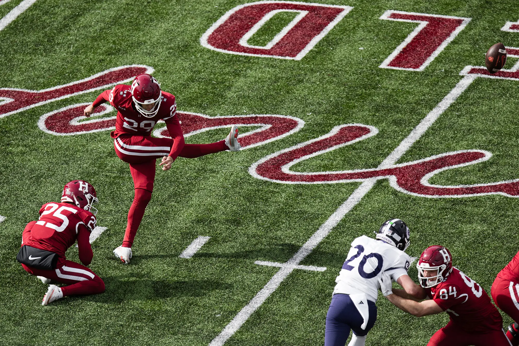 Harvard player #29, wearing a red uniform, kicks the ball with teammate #25 assisting while Harvard player #84 holds off Holy Cross player #20