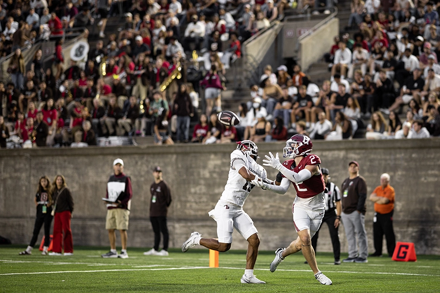 Two players battle for a pass mid-air with crowd watching intently in the stadium background.