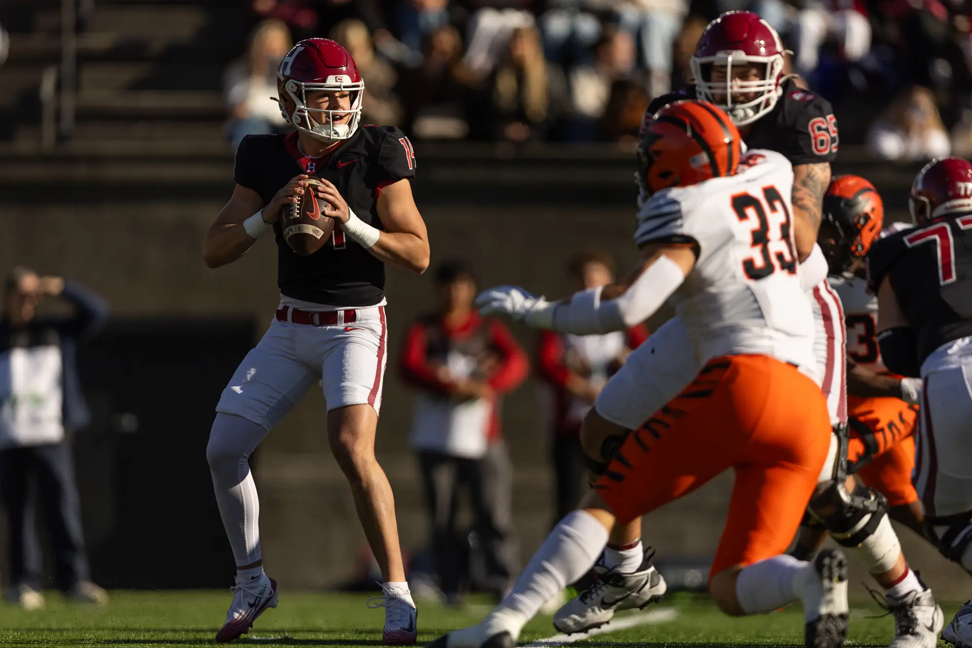 Harvard player #14, wearing a black uniform, prepares to throw the ball as Princeton player #33 wearing a white and orange uniform, is stopped by Harvard player #65