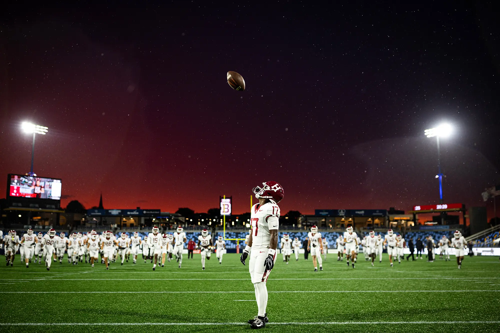 Harvard player #17, wearing a white uniform, watches as ball thrown in the air comes down against the night time sky with the team in a line across the field run towards player #17