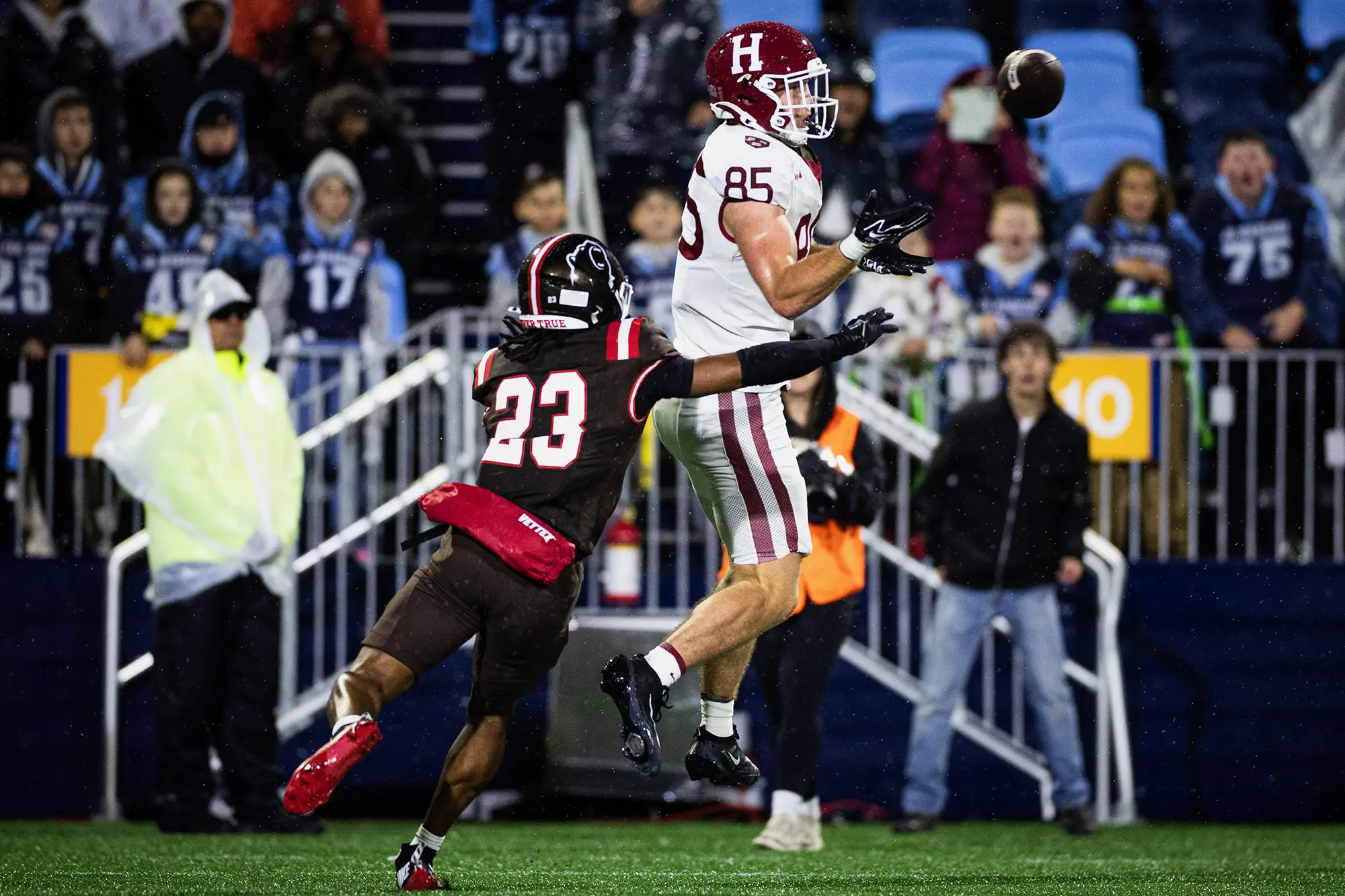 Harvard player #85 wearing a white uniform jumps in the air to catch an incoming ball while #23 from the opposing team, wearing a black uniform, attempts to intercept the catch.