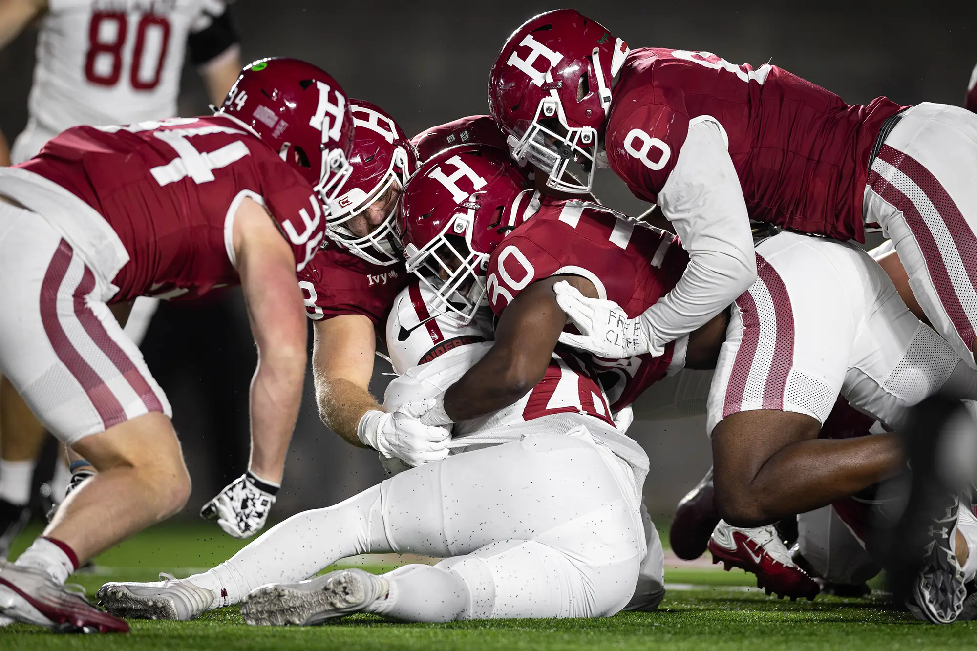 Harvard players wearing red uniforms collapse on a Colgate player wearing a white uniform