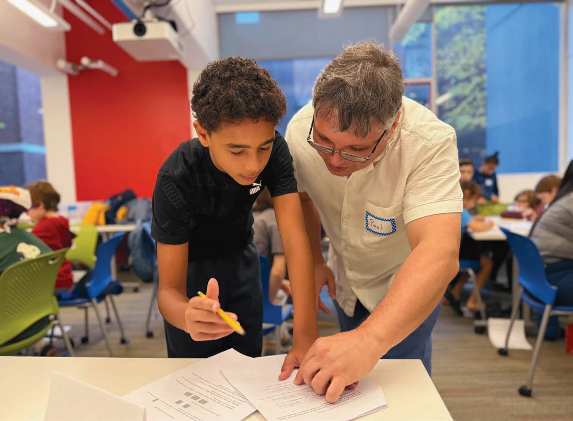 Adult helping a student with worksheets in a classroom filled with other children and educators.