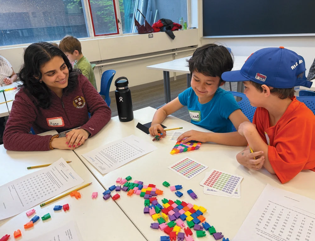 Three children and a tutor working with colorful math cubes and number grids at a classroom table.