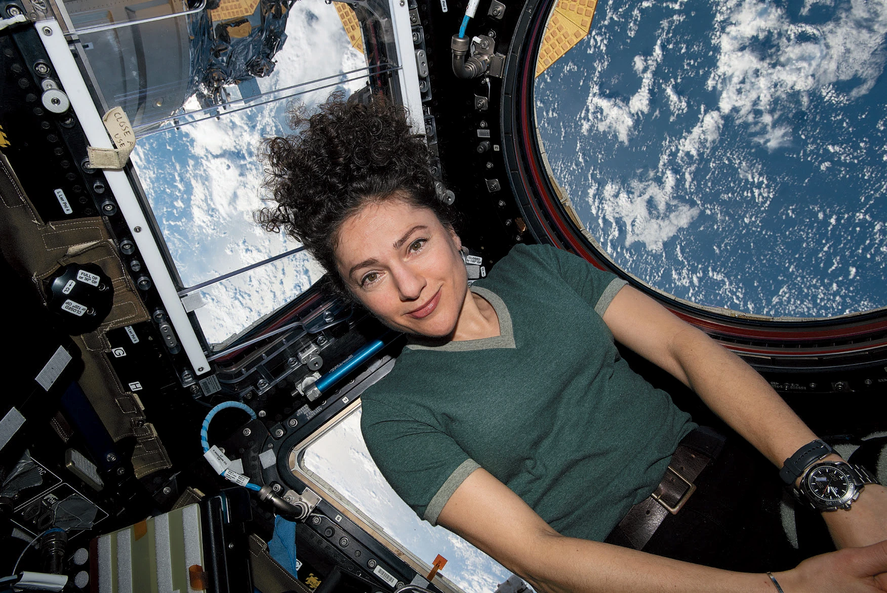 Jessica Meir inside a spacecraft looking at the camera with Earth visible through the window behind her.