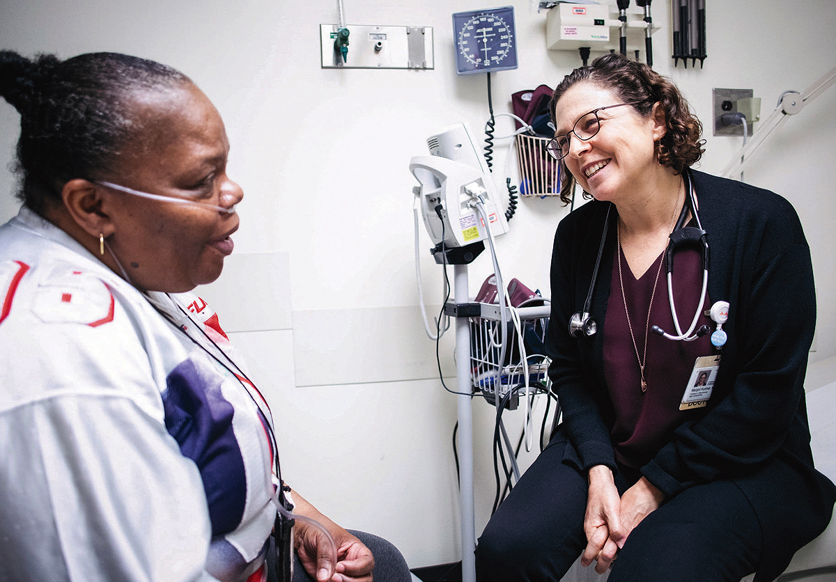 Seated in a clinical exam room, Kushel talks with a patient wearing an oxygen tube