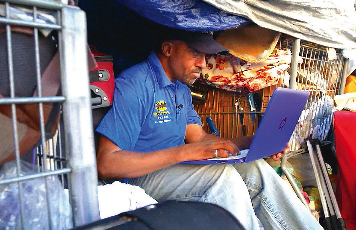 Pleasants wearing a blue shirt and jeans, sitting in his tent and typing on a laptop