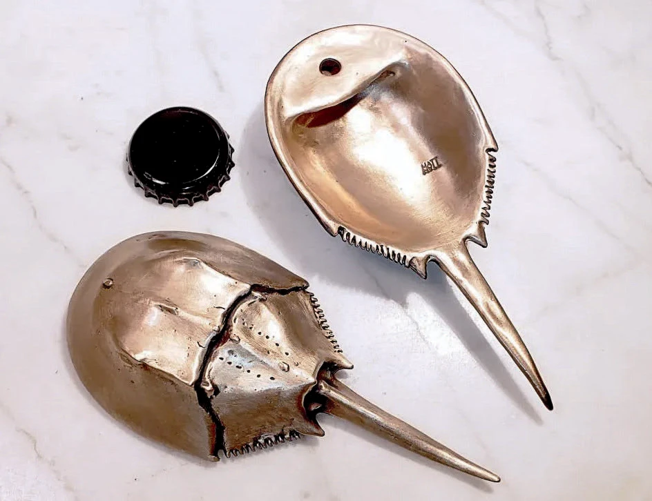 Two metal bottle openers shaped like horseshoe crabs beside a bottle cap for scale.
