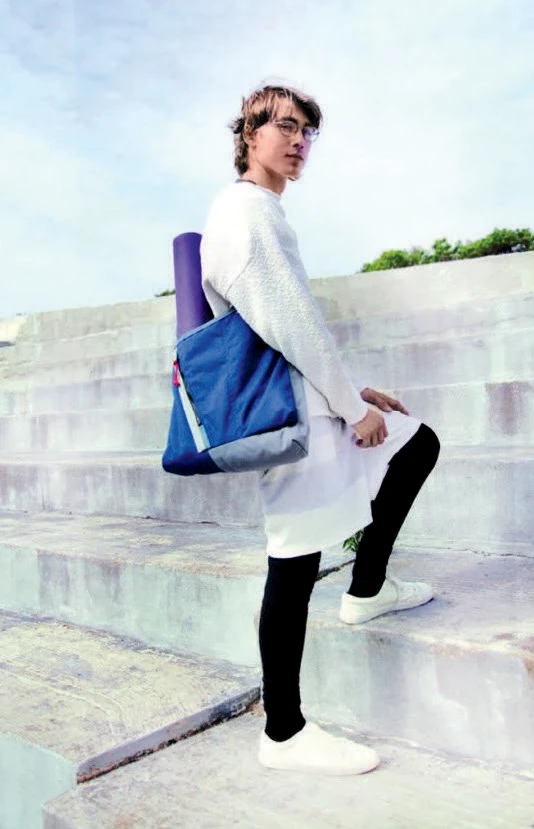 Person in layered clothing on stairs carrying a yoga mat in a blue tote bag.