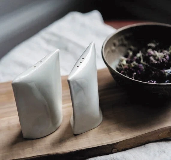 White modern salt and pepper shakers beside a ceramic bowl on a wooden board.