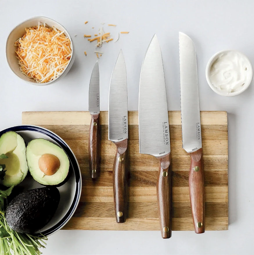 Set of four kitchen knives on a wooden board with shredded cheese, avocado, and sour cream nearby.