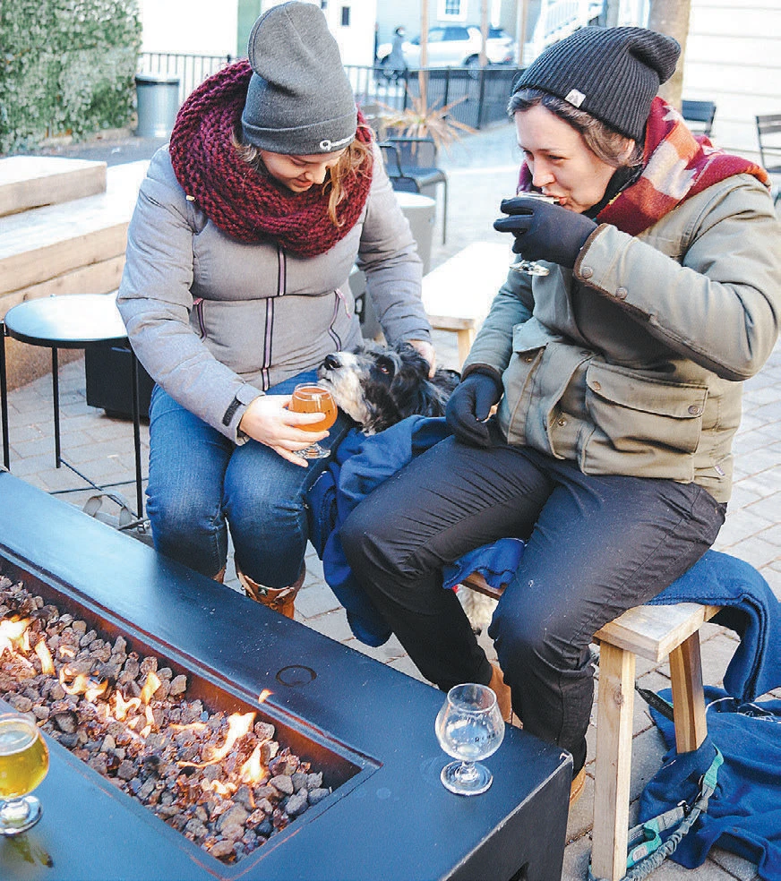 Two people in winter clothing drinking by a fire pit, petting a black and white dog.