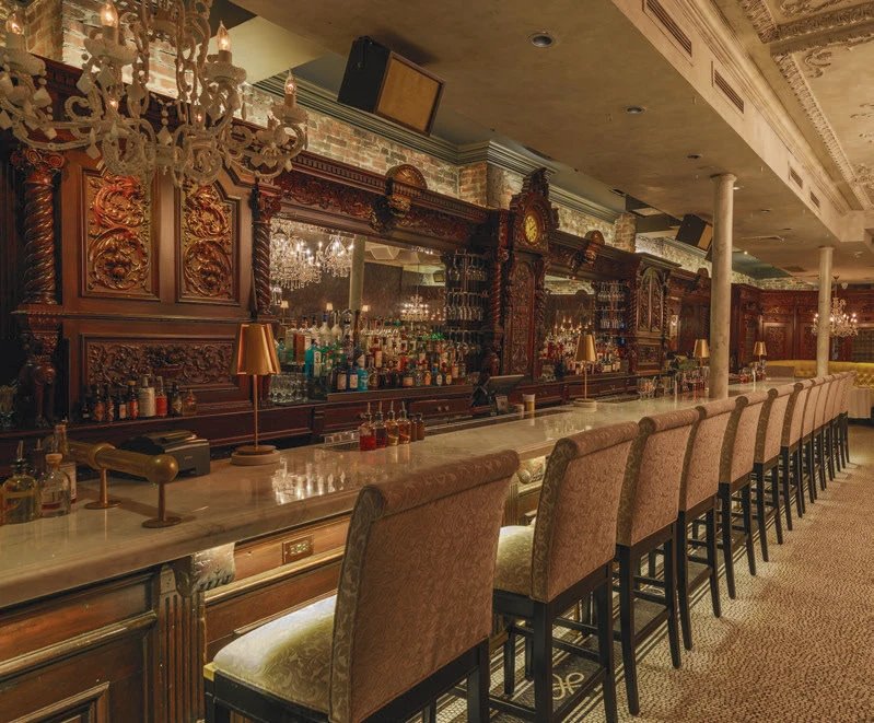 Ornate bar with carved wood, chandeliers, a marble counter, and rows of high-back chairs.