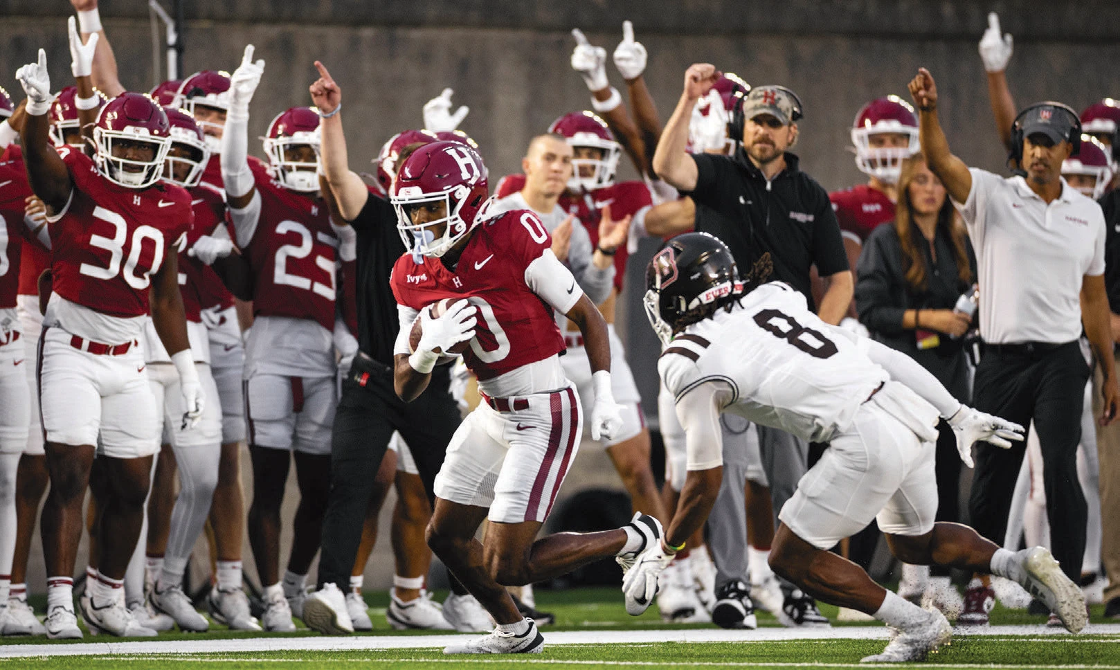 Harvard football player runs with the ball past a defender as teammates and coaches cheer on the sideline.
