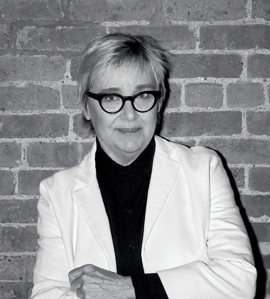Black and white portrait of Julie Bargmann in glasses and light blazer standing against a brick wall.