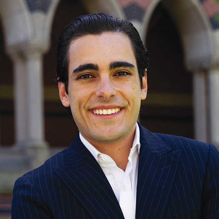 Michael Oved, smiling and wearing a dark pinstripe suit and white shirt standing in front of an arched building.
