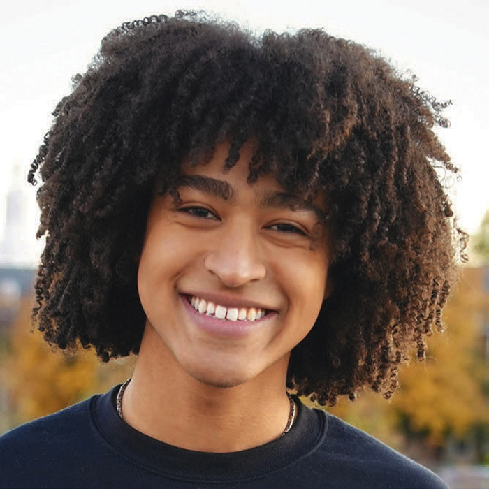 Smiling person with curly hair wearing a dark shirt, standing outdoors with blurred autumn trees in the background.