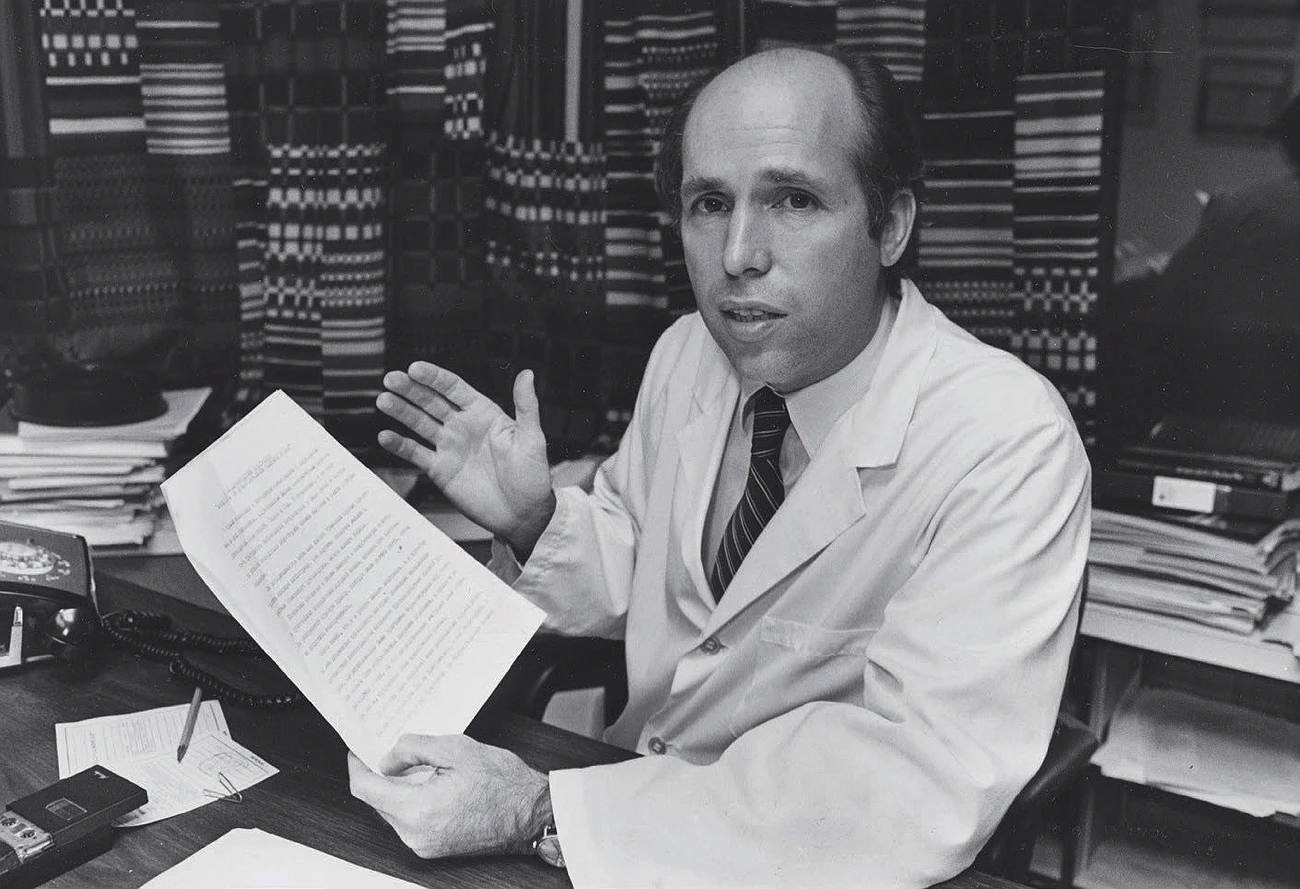 James Muller in white lab coat at desk holding letter and gesturing with hand.