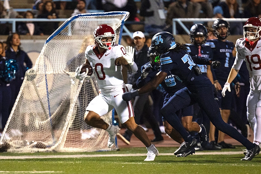 Harvard player runs with ball while fending off a tackle from Columbia defender near the sideline.