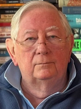 Older man with glasses and light hair, neutral expression, bookshelves behind