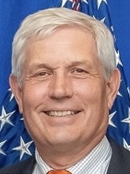 Older man in suit with gray hair, smiling, U.S. flag backdrop