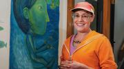 A woman in an orange sweater and cap stands in front of a large blue and green painting of a water-skiing woman.