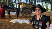 A woman with a backpack and a cup of coffee in a forest with fake snow and a small horse-drawn wagon. 