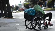 A man in a wheelchair with a rolled-up sleeping bag strapped to the back