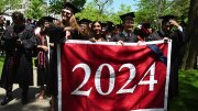 Graduates wearing black caps and gowns processing through Harvard Yard while holding the Class of 2024 banner