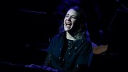 Madeline Benson singing at the piano, her face lit up on an otherwise dark stage