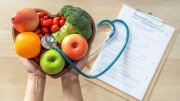 Hands holding a heart-shaped bowl of fruits and vegetables with a stethoscope, clipboard with medical records in the background.