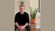 Richard Beck sitting indoors beside a potted plant, hands clasped, looking at the camera.
