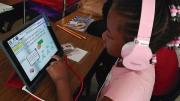 A young girl with pink headphones uses a tablet for an educational reading game at a classroom desk.