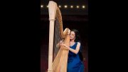 Elisabeth Remy Johnson playing a harp on stage, wearing a blue dress, with stage lights softly glowing in the background.